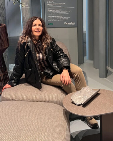 Projektant i architekt wnętrz Anna Buczny, siedzi na sofie firmy Varaschin. Projektantka ubrana jest w czarną skórę, sofa jest w kolorach ziemi, podobnie jak stolik stojący przy sofie.