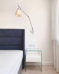 Ciemne wezgłowie łóżka obok, którego stoi szklana szafeczka nocna firmy Fiam Italia, kontrastuje z jasnymi ścianami. Listwy przypodłogowe pomalowane w kolorze ścian. Podłoga panelowa w jasnym odcieniu drewna jest ciepłym akcentem wnętrza utrzymanego w stylu skandynawskim. Architekt wnętrz Anna Buczny zrealizowała aranżację sypialni w formule pod klucz.