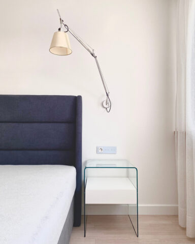 Ciemne wezgłowie łóżka obok, którego stoi szklana szafeczka nocna firmy Fiam Italia, kontrastuje z jasnymi ścianami. Listwy przypodłogowe pomalowane w kolorze ścian. Podłoga panelowa w jasnym odcieniu drewna jest ciepłym akcentem wnętrza utrzymanego w stylu skandynawskim. Architekt wnętrz Anna Buczny zrealizowała aranżację sypialni w formule pod klucz.