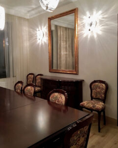 Jadalnia w klasycznym stylu, z ciemnymi meblami. Krzesła tapicerowane wzorzystą tkaniną, stoją przy stole i przy ścianie - po obu stronach kredensu. Drewniana, dębowa podłoga kontrastuje z ciężkim, drewnianym, ciemnym stołem. Duże lustro w złotej ramie, zawieszone nad kredensem, optycznie powiększa pomieszczenie. Dekoracyjne, kryształowe kinkiety zamontowane po obu stronach lustra i wiszące lampy nad stołem, pochodzą z tej samej kolekcji lamp Schonbek. Dekoracyjne tkaniny w oknach, zmiękczają ściany i nawiązują do pastelowej kolorystyki ścian. Wnętrze zaprojektowała i wykonała pod klucz architekt wnętrz Anna Buczny.