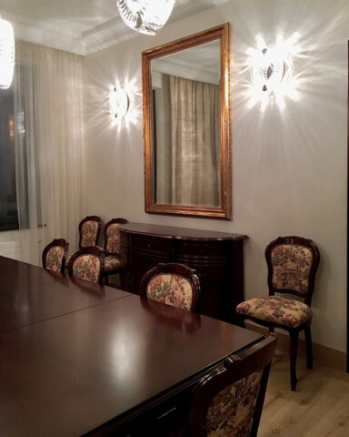 Jadalnia w klasycznym stylu, z ciemnymi meblami. Krzesła tapicerowane wzorzystą tkaniną, stoją przy stole i przy ścianie - po obu stronach kredensu. Drewniana, dębowa podłoga kontrastuje z ciężkim, drewnianym, ciemnym stołem. Duże lustro w złotej ramie, zawieszone nad kredensem, optycznie powiększa pomieszczenie. Dekoracyjne, kryształowe kinkiety zamontowane po obu stronach lustra i wiszące lampy nad stołem, pochodzą z tej samej kolekcji lamp Schonbek. Dekoracyjne tkaniny w oknach, zmiękczają ściany i nawiązują do pastelowej kolorystyki ścian. Wnętrze zaprojektowała i wykonała pod klucz architekt wnętrz Anna Buczny.